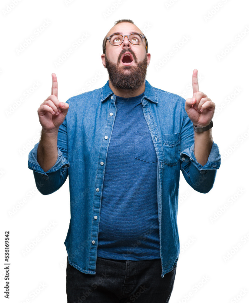 Young caucasian hipster man wearing glasses over isolated background amazed and surprised looking up and pointing with fingers and raised arms.