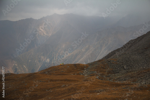 A mountain gorge with huge slopes, a distant slope in the fog, the front slope is rocky, there is a small house, autumn is in the mountains, the sun shines faintly through the fog