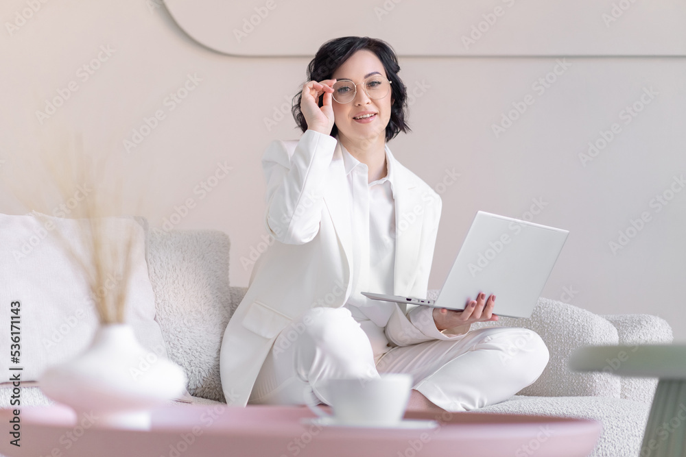 Business happy middle aged woman sitting on sofa in bright workspace. He holds glasses in one hand and a laptop in the other. Blurred foreground. Business happy middle aged woman sitting on sofa in bright workspace. He holds glasses in one hand and a laptop in the other. Blurred foreground.