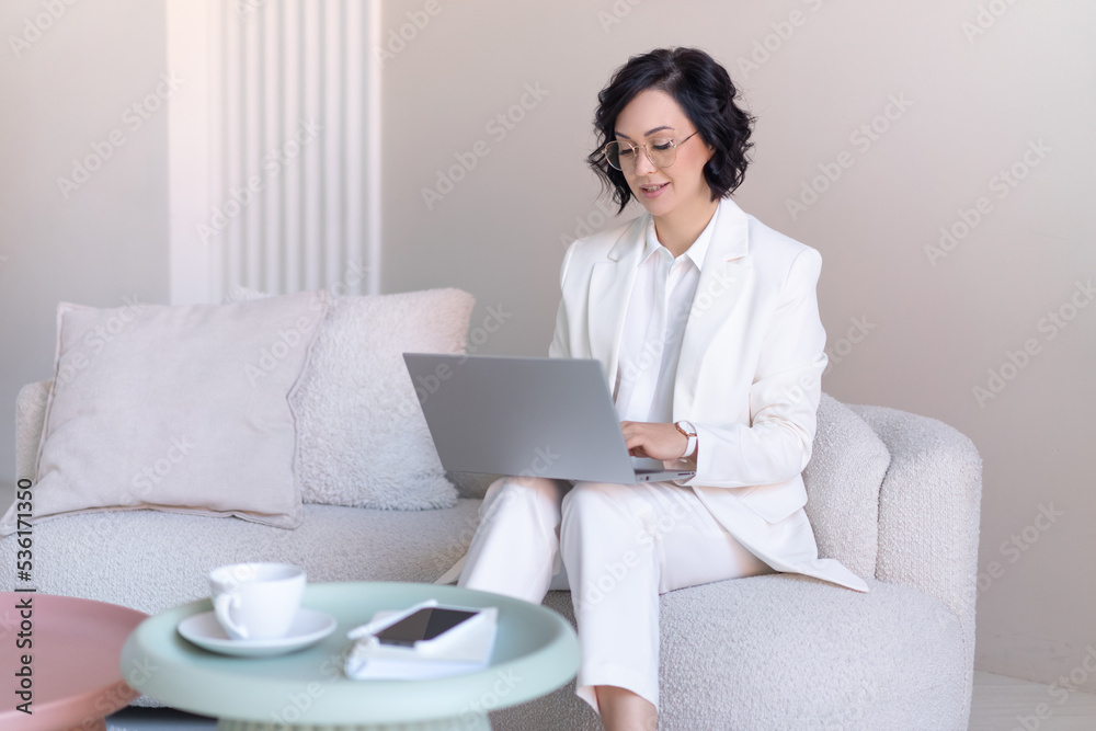 Middle-aged business woman is typing on a laptop keyboard while sitting on a sofa in her home office. Blurred foreground. Middle-aged business woman is typing on a laptop keyboard while sitting on a sofa in her home office. Blurred foreground.