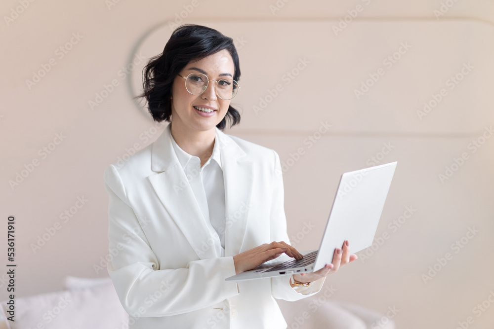 Portrait of smiling middle-aged brunette woman in white jacket. With silver laptop in hand. Looks into camera. With copy space. Portrait of smiling middle-aged brunette woman in white jacket. With silver laptop in hand. Looks into camera. With copy space.