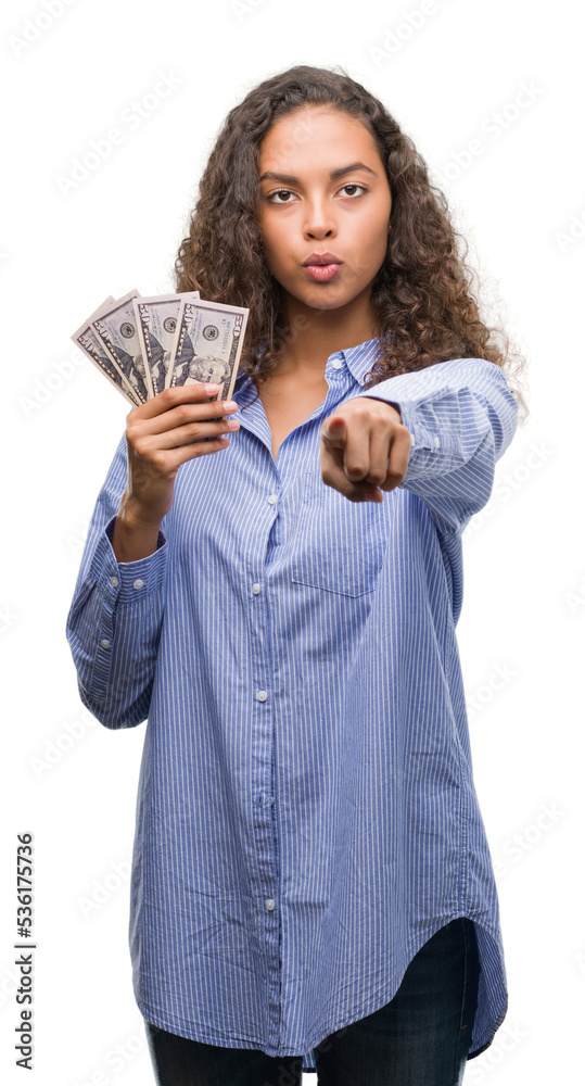 Young hispanic woman holding dollars pointing with finger to the camera and to you, hand sign, positive and confident gesture from the front Young hispanic woman holding dollars pointing with finger to the camera and to you, hand sign, positive and confident gesture from the front