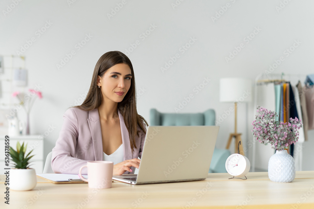 Young smiling business woman in a pink jacket sits at a table and works on a laptop. Looks into the camera. Cozy light minimalist office. Distant work. Copy space. Young smiling business woman in a pink jacket sits at a table and works on a laptop. Looks into the camera. Cozy light minimalist office. Distant work. Copy space.