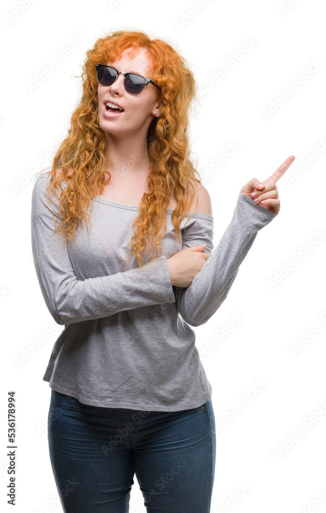 Young redhead woman wearing sunglasses very happy pointing with hand and finger to the side Young redhead woman wearing sunglasses very happy pointing with hand and finger to the side