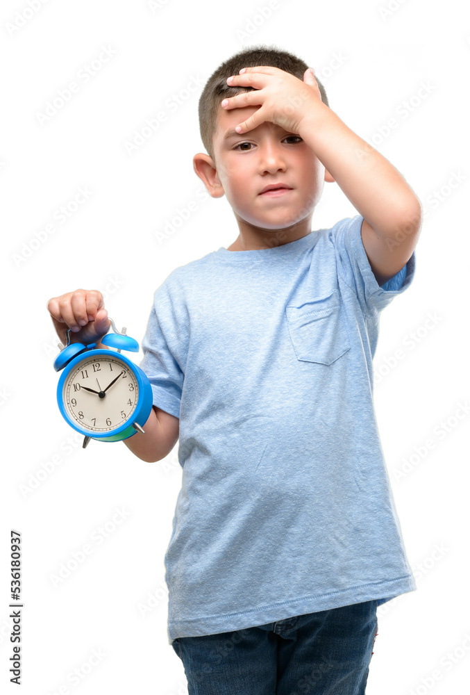 Little child holding alarm clock stressed with hand on head, shocked