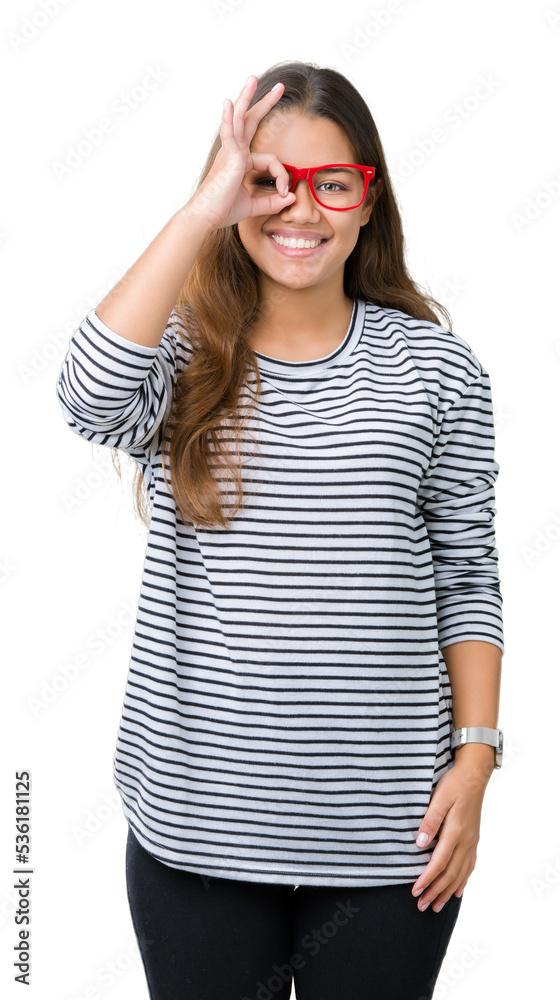 Young beautiful brunette woman wearing stripes sweater and red glasses over isolated background doing ok gesture with hand smiling, eye looking through fingers with happy face. Young beautiful brunette woman wearing stripes sweater and red glasses over isolated background doing ok gesture with hand smiling, eye looking through fingers with happy face.