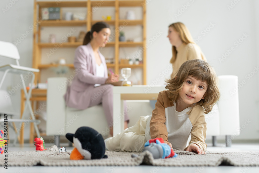 Male child is playing on floor with his toys. Mom communicates with child psychologist sitting on couch. Office of child psychologist. mental health.