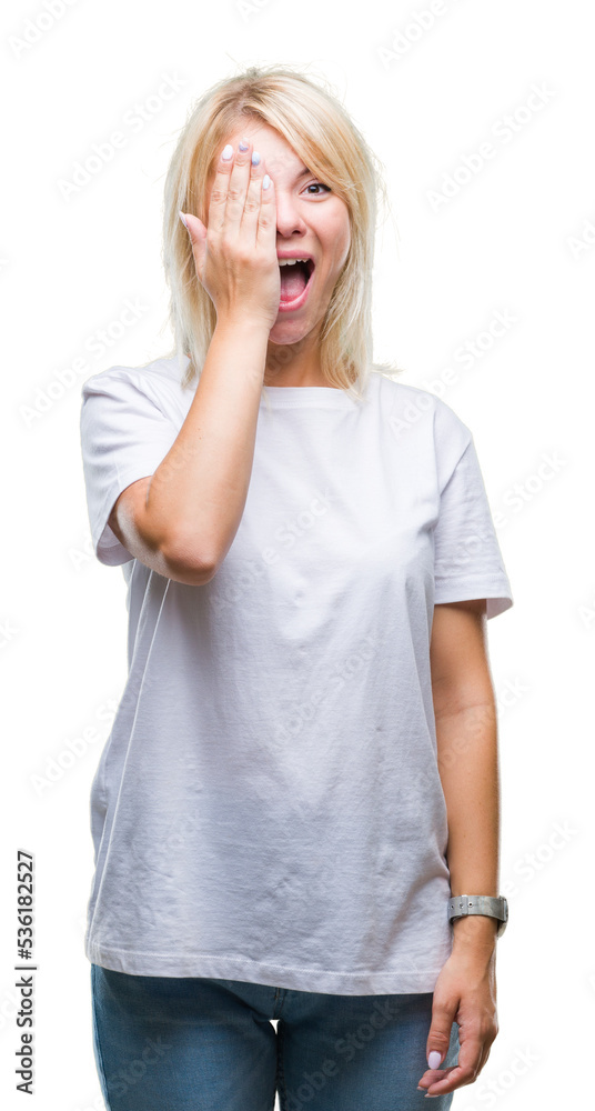 Young beautiful blonde woman wearing white t-shirt over isolated background covering one eye with hand with confident smile on face and surprise emotion.