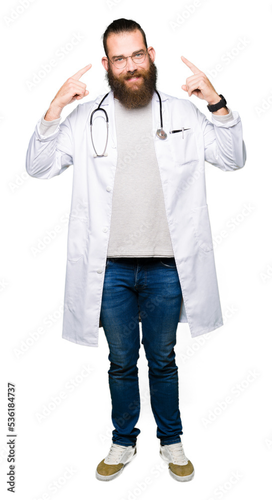 Young blond doctor man with beard wearing medical coat Smiling pointing to head with both hands finger, great idea or thought, good memory Young blond doctor man with beard wearing medical coat Smiling pointing to head with both hands finger, great idea or thought, good memory