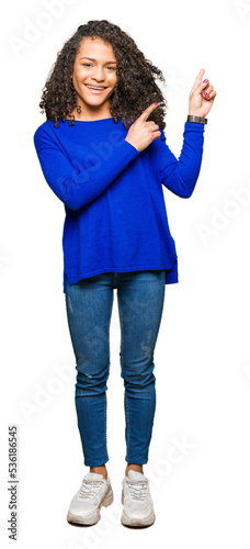 Young beautiful woman with curly hair wearing winter sweater smiling and looking at the camera pointing with two hands and fingers to the side.