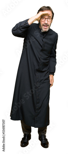 Fotografie, Obraz Middle age priest man wearing catholic robe very happy and smiling looking far away with hand over head