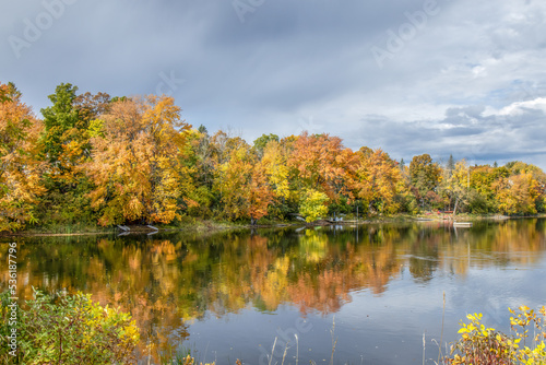View down a calm river in autumn, fall foliage on riverbank reflected in water, docks, canoes, dark clouds, nobody