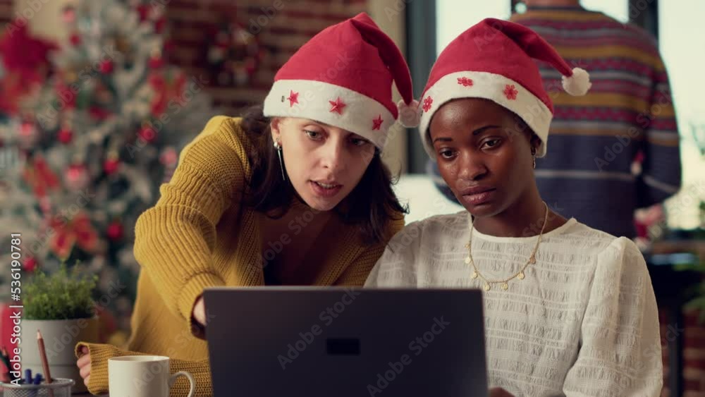 Multiethnic team of coworkers planning report on laptop, using research data in office with christmas tree and ornaments. Diverse people with santa hats celebrating xmas winter holiday. Multiethnic team of coworkers planning report on laptop, using research data in office with christmas tree and ornaments. Diverse people with santa hats celebrating xmas winter holiday.
