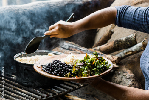 Hands of an unrecognizable woman serving a plate of rice with beans and salad in a traditional kitchen.