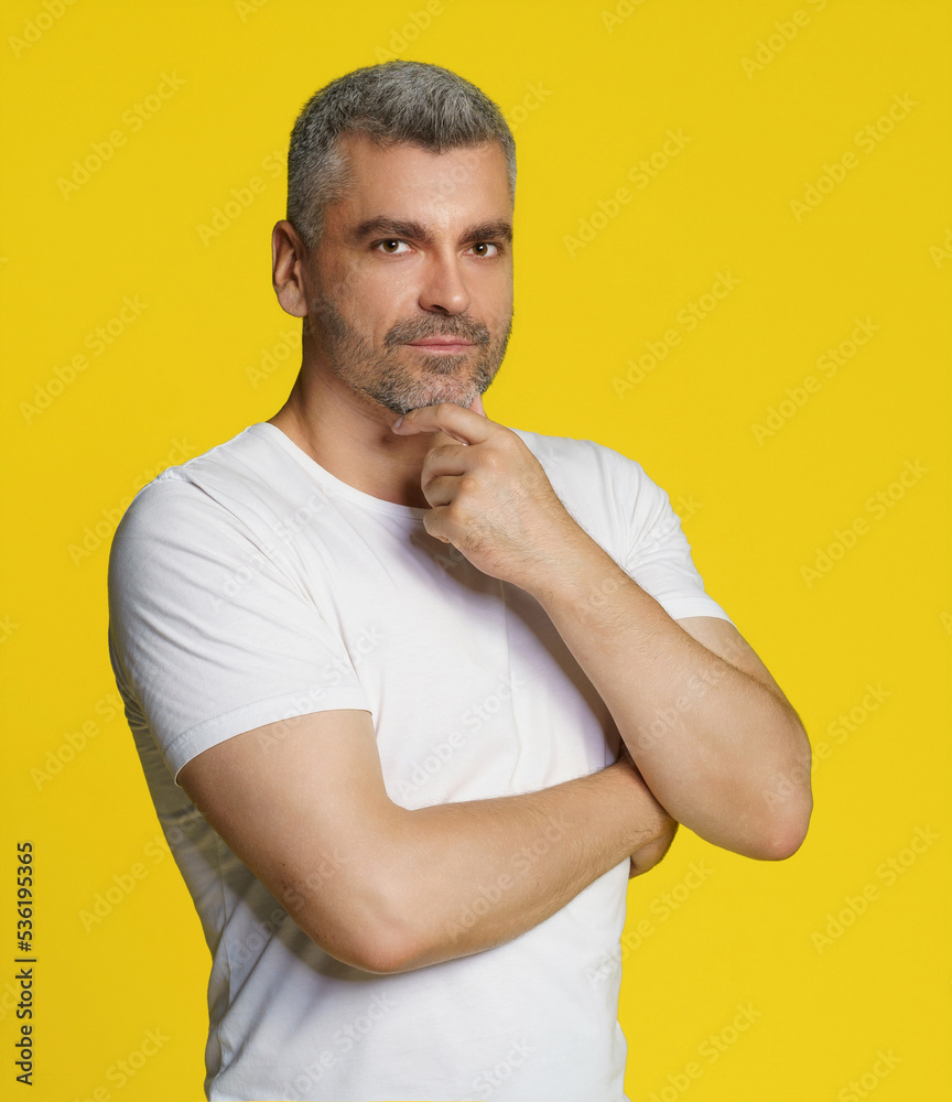 Middle aged grey hair athletic man posing with hands crossed touching chin wearing white t-shirt isolated on yellow background. Thoughtful fit, muscular middle aged man Middle aged grey hair athletic man posing with hands crossed touching chin wearing white t-shirt isolated on yellow background. Thoughtful fit, muscular middle aged man