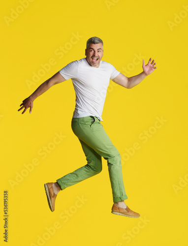 Handsome man with grey hair jump in the air turned sideways wearing white t-shirt and green jeans isolated on yellow background. Excited, happy fit, muscular middle aged man
