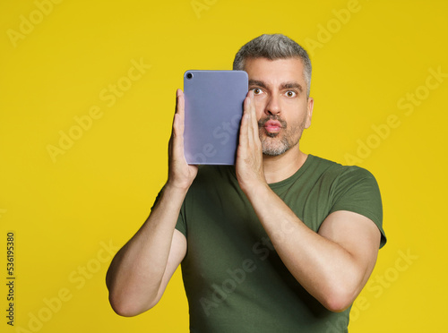 Handsome man send kisses holding digital tablet using dating app in hands looking at camera isolated on yellow background. Happy man with tablet. Online dating service. mobile app advertisement