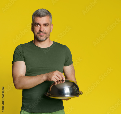 Handsome man with grey hair hold metallic cloche serving food isolated on yellow background. Romantic dinner at home man surprises girlfriend with cooking skills. Service, restaurant, horeca concept photo