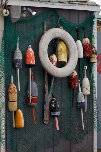 A collection of old colorful buoys, fishing net, and life preserver are hanging from gray wall outside of fishing shack on an overcast day.