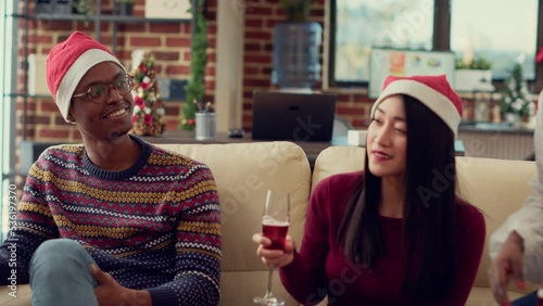 Diverse employees drinking glasses of wine at christmas party in office decoratd with festive ornaments. Serving alcoholic drinks at winter holiday festivity, wearing santa hats. Handheld shot. photo