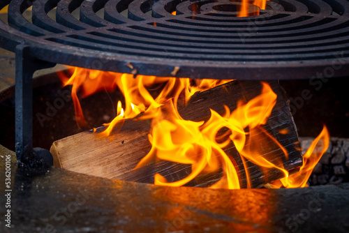 A wooden log burns brightly in a wood-fired grill. Circle wood-fired grill with steel grate for outdoor cooking