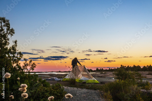 Obraz na plátne A young man sets up a small tent on a rocky expanse with the sun setting in the background