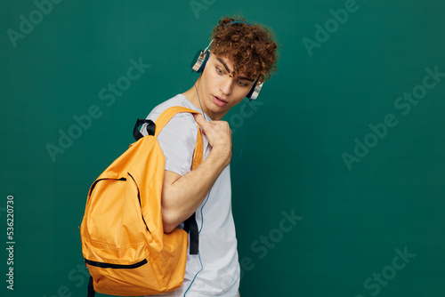 horizontal portrait of a handsome man with curly hair standing on a green background in a gray T-shirt, listening to music with headphones, looking at his bright backpack