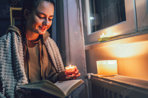 Teenage girl sits under blanket near heating radiator with candles and read book .Rising costs in private households for gas bill due to inflation and war, Energy crisis