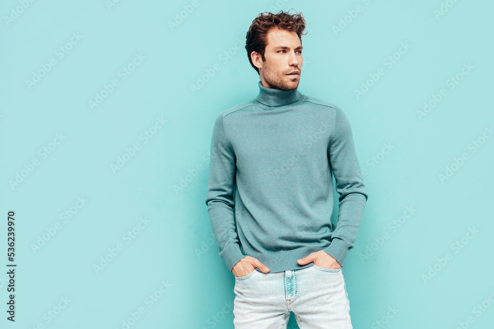 Portrait of handsome confident model. Sexy stylish man dressed in sweater and jeans. Fashion hipster male with curly hairstyle posing near blue wall in studio. Isolated Portrait of handsome confident model. Sexy stylish man dressed in sweater and jeans. Fashion hipster male with curly hairstyle posing near blue wall in studio. Isolated