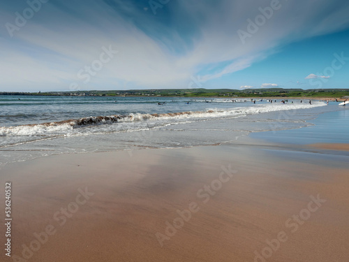 Yellow sand and blue ocean and sky. People swim in the background and surf on board. Lahinch beach in county Clare, Ireland. Warm sunny day. Irish landscape.
