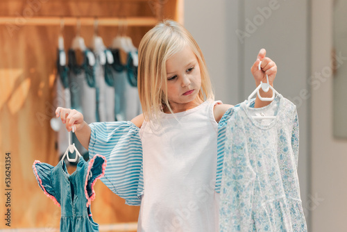 Canvas Print Child, fashion and shopping while choosing kids clothing in a shop, store or boutique with a cute girl holding dresses thinking or picking color choice