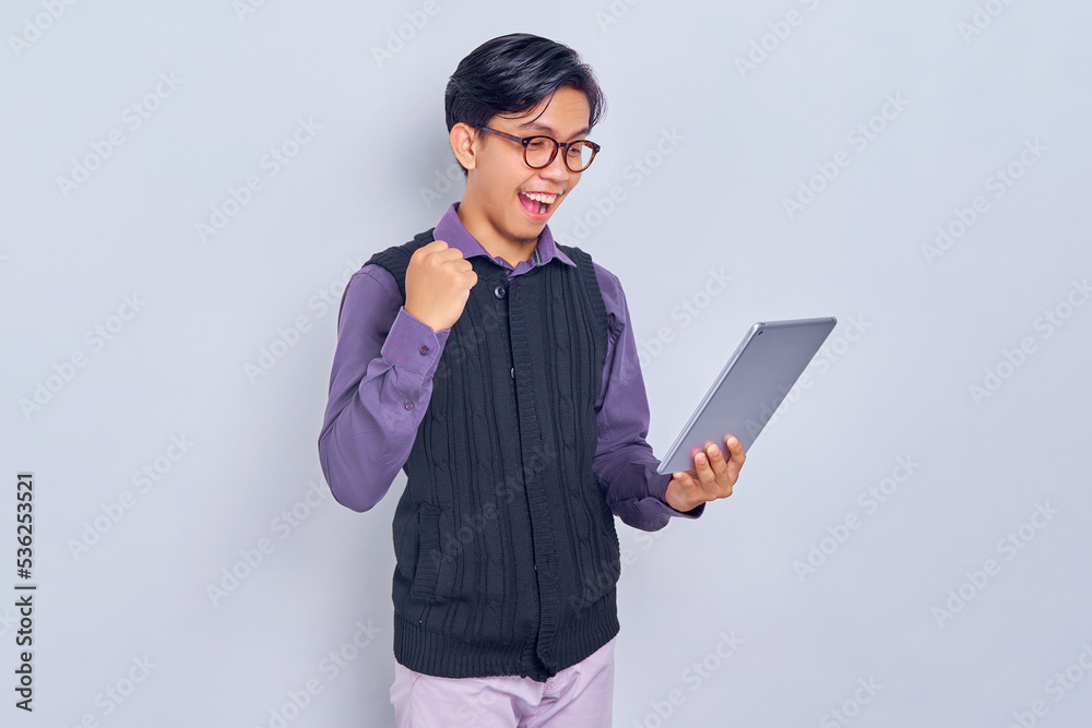 Smiling young Asian man in casual shirt and vest holding tablet pc computer and making winner gesture isolated on white background. People lifestyle concept Smiling young Asian man in casual shirt and vest holding tablet pc computer and making winner gesture isolated on white background. People lifestyle concept
