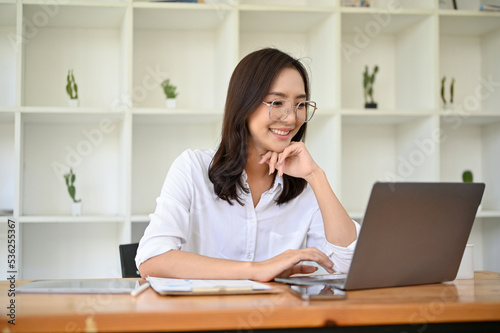 Attractive Asian businesswoman in eyeglasses looking at laptop screen, hand on chin