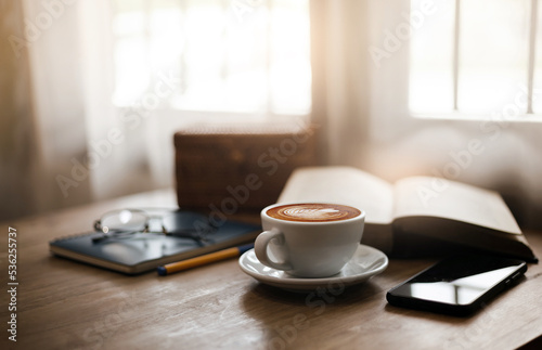 Close up view, Latte coffee in white cup and smart phone on wooden table near bright window. blurred background with book, eyeglasses on blue note book and pen, sepia color tone