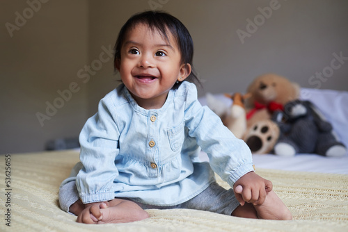 Baby, down syndrome and special needs child happy with a smile sitting on a bed to relax, be curious and playful in bedroom at home. Cheerful girl with intellectual disability and developmental delay photo