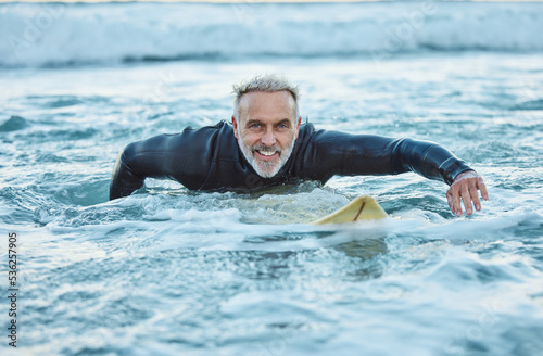 Senior man, water and sports surfer on a beach with smile training for fitness and health or hobby in the outdoors. Elderly male paddling on surfboard for healthy exercise in the ocean waves at sea