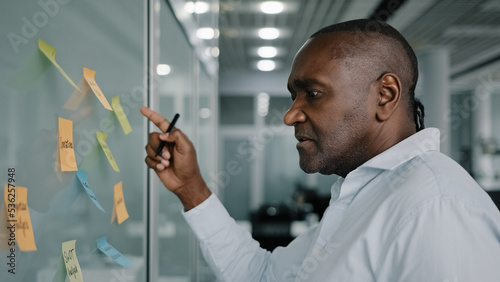 African businessman executive content manager develop strategy plan manage corporate work tasks read sticky notes glued to board write ideas project points on memo stickers planning goals brainstorm