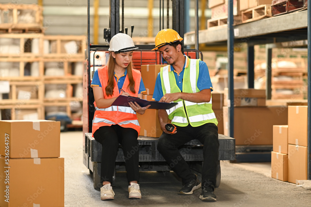 Two warehouse workers wearing safety hard hat and vest sitting on forklift truck in retail warehouse checking newly arrived goods