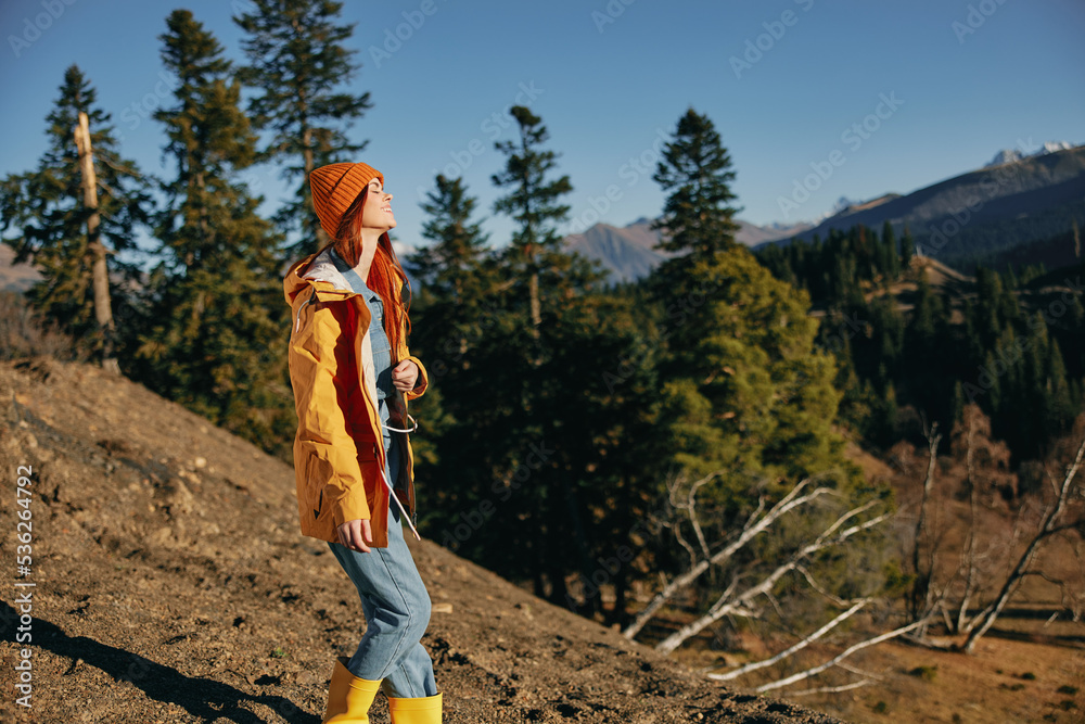 Naklejka premium Woman traveling to the mountains in the fall on a nature hike smile and happiness in a yellow cape with red hair full-length stands against the backdrop of trees and mountains