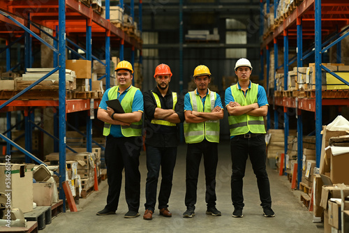 Group of warehouse workers in hardhat and reflective jacket standing with arm crossed between rows of tall shelves at retail warehouse