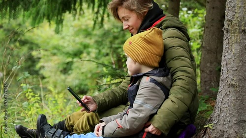 Joyful happy traveling mother and son using smartphone mobile resting in mountains forest near the lake enjoying beautiful autumn nature. Internet connecting 5g. Young family lifestyle.