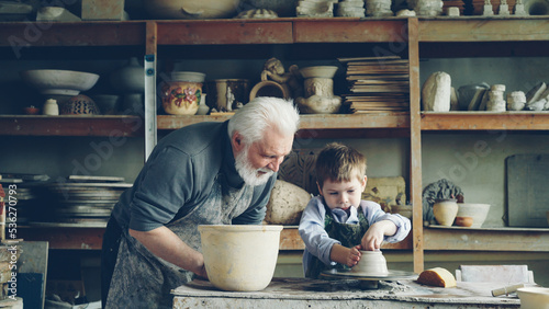 Fotografija Curious little boy is learning pottery from his experienced grandfather in small home studio