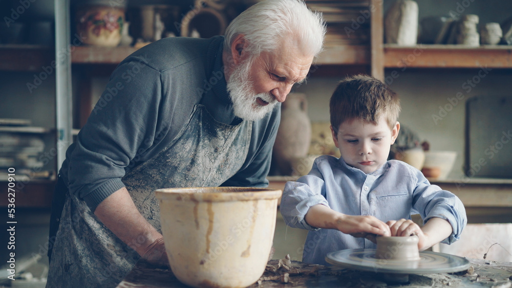 Concentrated young boy is molding clay into ceramic pot on spinning throwing wheel and his loving experienced grandfather is talking to him. Pottery and family tradition concept.