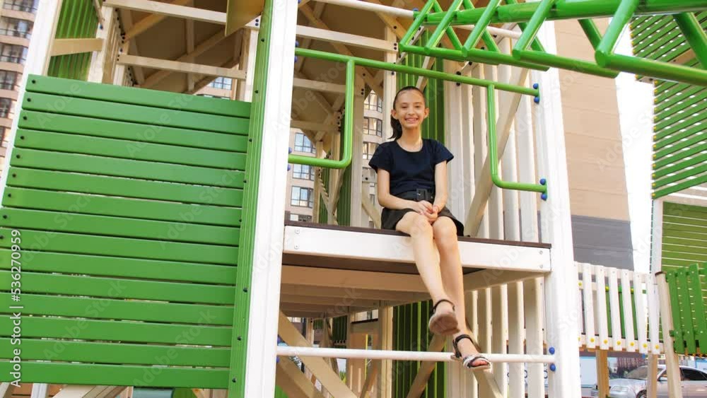 A happy 8-year-old girl sits in a house on the playground and shakes her legs. The happiness of children. Playgrounds in the city. A happy 8-year-old girl sits in a house on the playground and shakes her legs. The happiness of children. Playgrounds in the city.