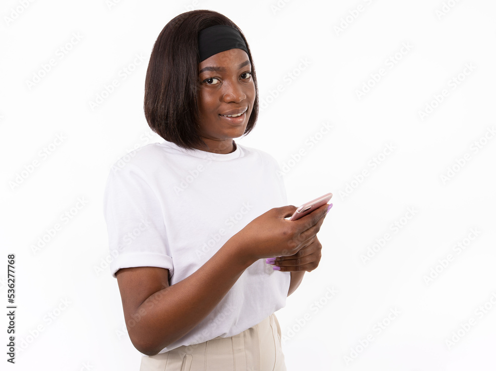 Naklejka premium Portrait of happy young woman using smartphone. African American lady wearing white T-shirt and headband looking at camera and smiling against white background. Mobile technology concept
