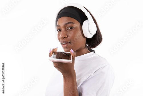 Portrait of happy young woman listening to music on smartphone. African American lady wearing white T-shirt and headband recording audio message in heaphones and looking at camera. Mobile app concept