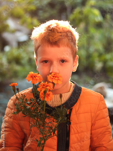 Red-haired teen boy in yellow clothes looking at camera sniffing a flower. A child with orange flowers near his face on natural background at sunset