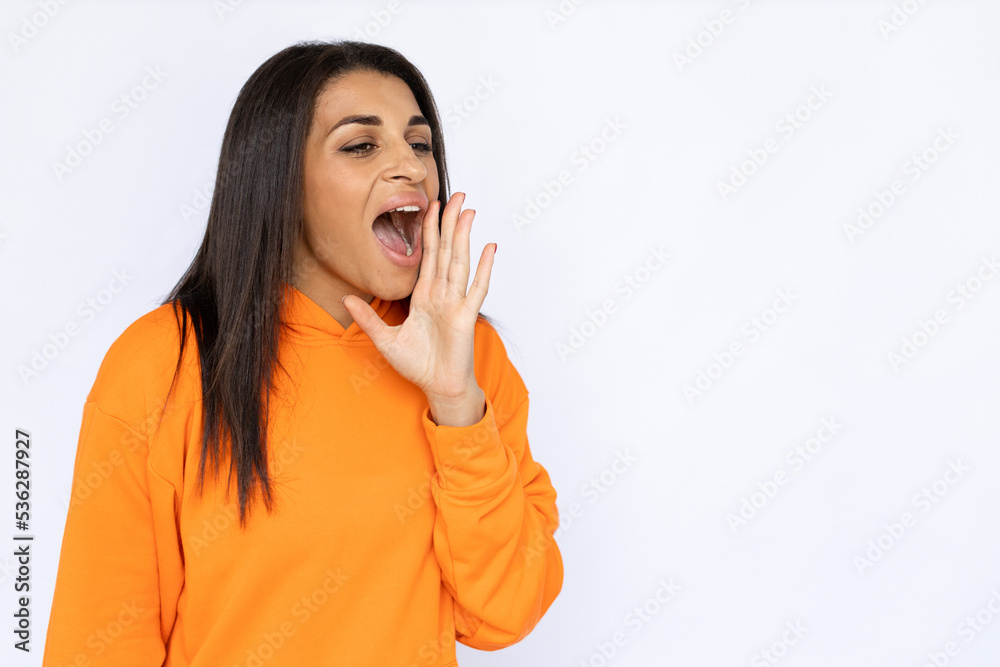 Fototapeta premium Portrait of screaming Latin American woman. Happy young model with long hair in orange hoodie looking away, shouting with hand near mouth. Studio shot, announcement concept.