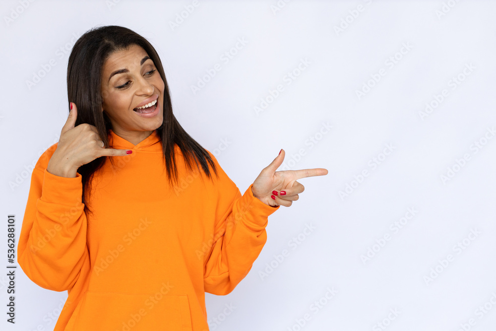 Portrait of happy Hispanic woman pointing and doing calling sign. Cheerful young model with long hair in orange hoodie looking away, smiling and showing ads. Advertisement concept. Portrait of happy Hispanic woman pointing and doing calling sign. Cheerful young model with long hair in orange hoodie looking away, smiling and showing ads. Advertisement concept.