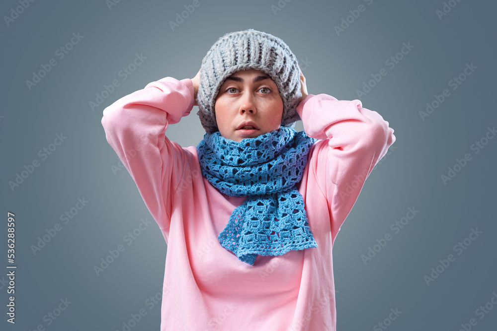 Illness and flu. Young tired woman in hat and scarf is warming herself and cough. Gray background. The concept of energy crisis and winter season Illness and flu. Young tired woman in hat and scarf is warming herself and cough. Gray background. The concept of energy crisis and winter season